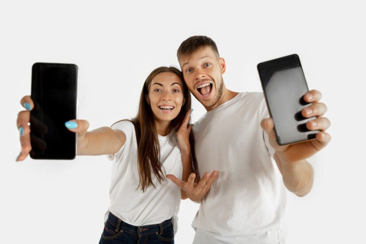 Um casal está segurando um celular cada um. Eles estão felizes e apontam para alguns dos Melhores Celulares 2024