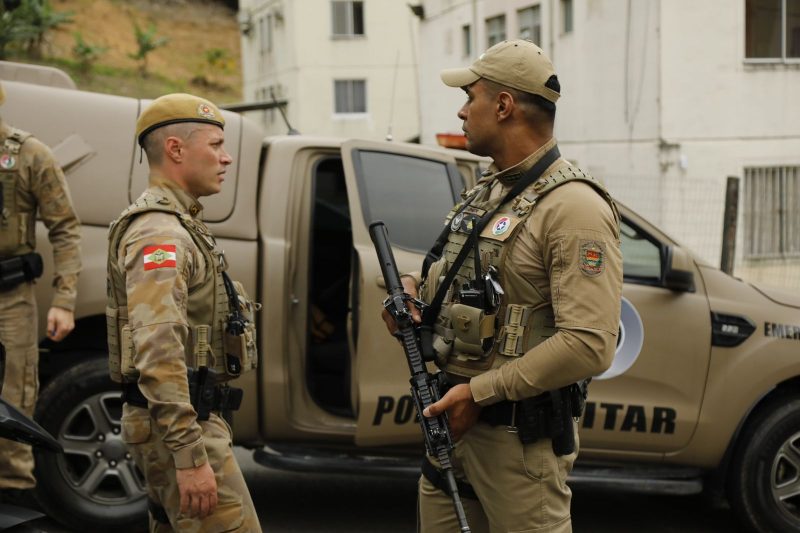 Foto mostra dois policiais militares e uma viatura da polícia.